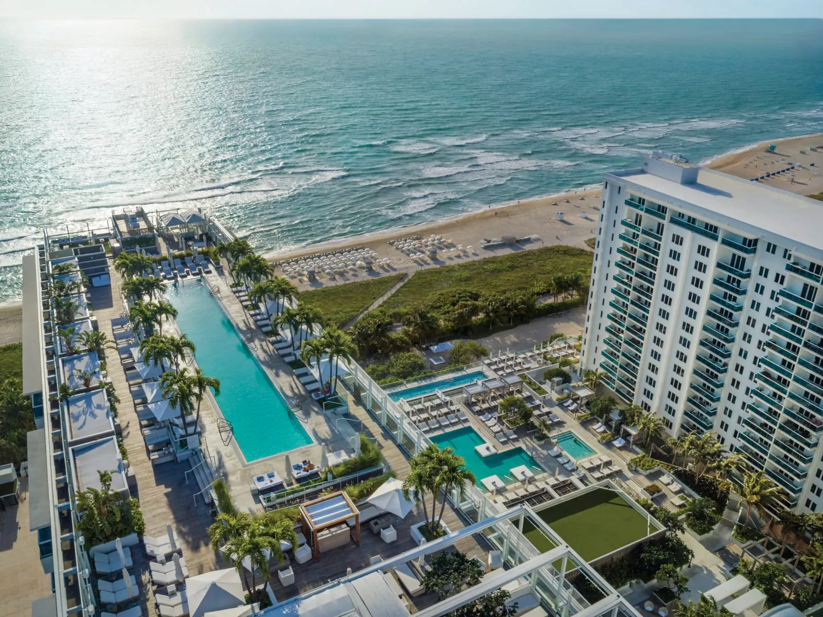 An aerial view down the property: a long lap pool runs parallel to the Atlantic, a second pool sits nearer the tower, and loungers line the beach beyond.