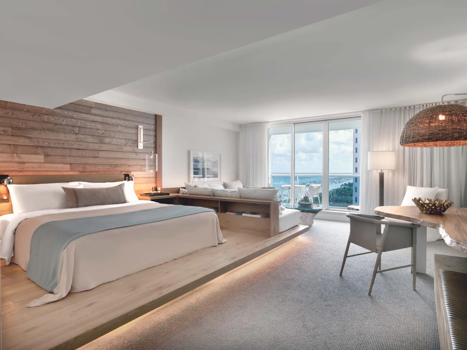 A bedroom on a raised wood platform, its headboard in weathered wood beside glass doors onto a balcony with a slice of ocean.