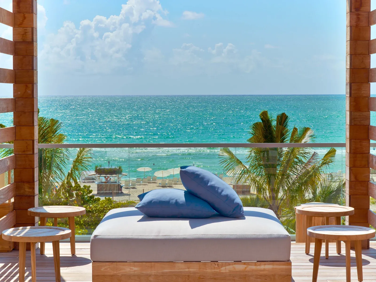 A shaded daybed on an oceanfront terrace, blue pillows facing the beach's loungers and umbrellas through the palms.