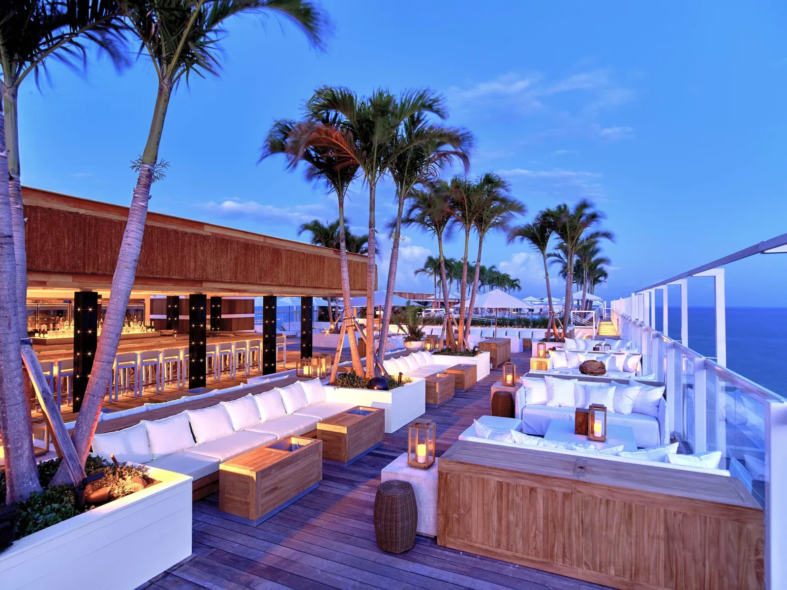 The 18th-floor adults-only rooftop at dusk, white sectionals and lantern candles under palms with the ocean past the glass rail.