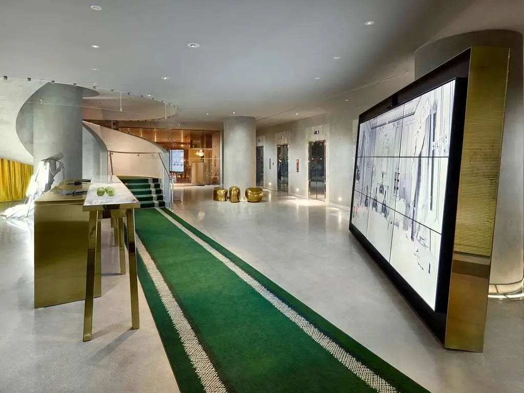 The lobby, raw concrete and a spiral stair beside a wall of brass-framed screens, a green racing-stripe runner underfoot.