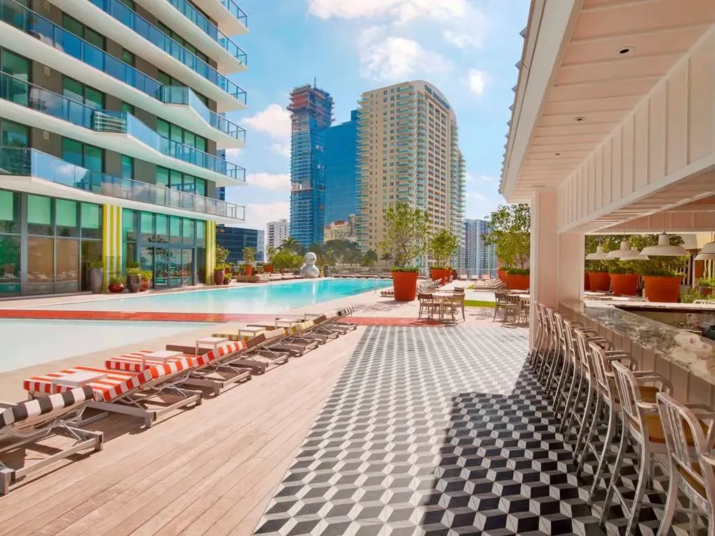 The rooftop deck's poolside bar on black-and-white checkerboard tile, striped loungers along the infinity pool with downtown Miami's towers behind.