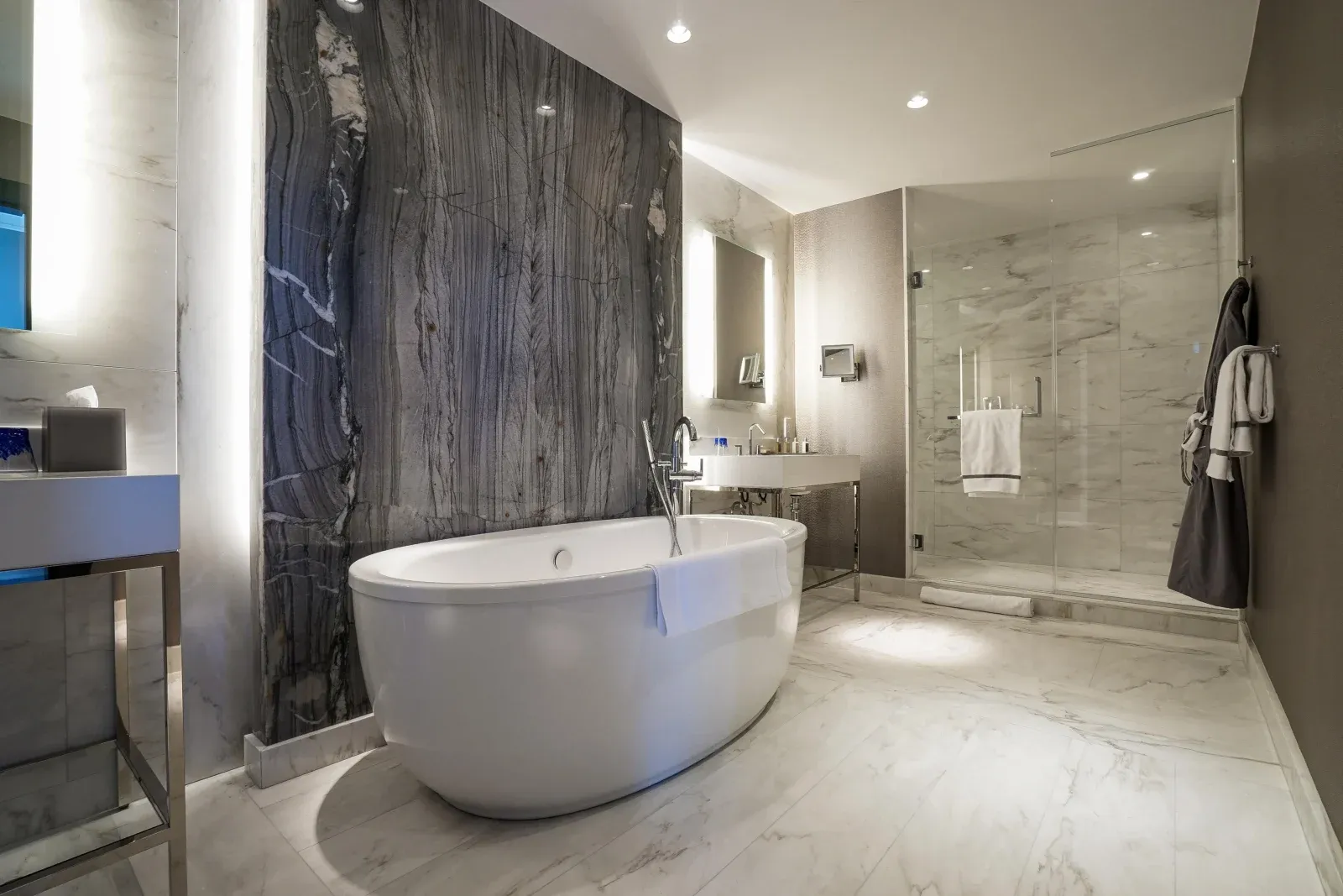 A freestanding oval tub against gray-veined marble, a glass shower alongside.