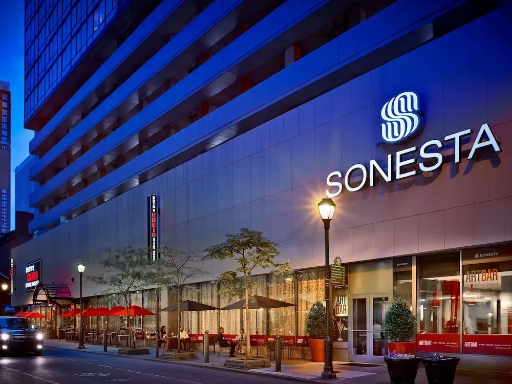 The Sonesta tower at dusk, red umbrellas and the lit street-level bar.