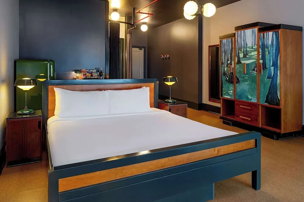 A teal and walnut bed frame beside a painted deco armoire and globe lights.